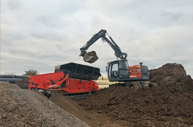 Bagger beim Beladen einer mobilen Brecheranlage mit Humus